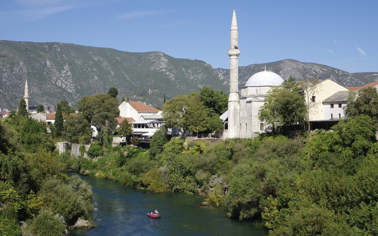 De groene rivier Neretva in Mostar met een moskee en minaret tussen de bomen en bergen op de achtergrond