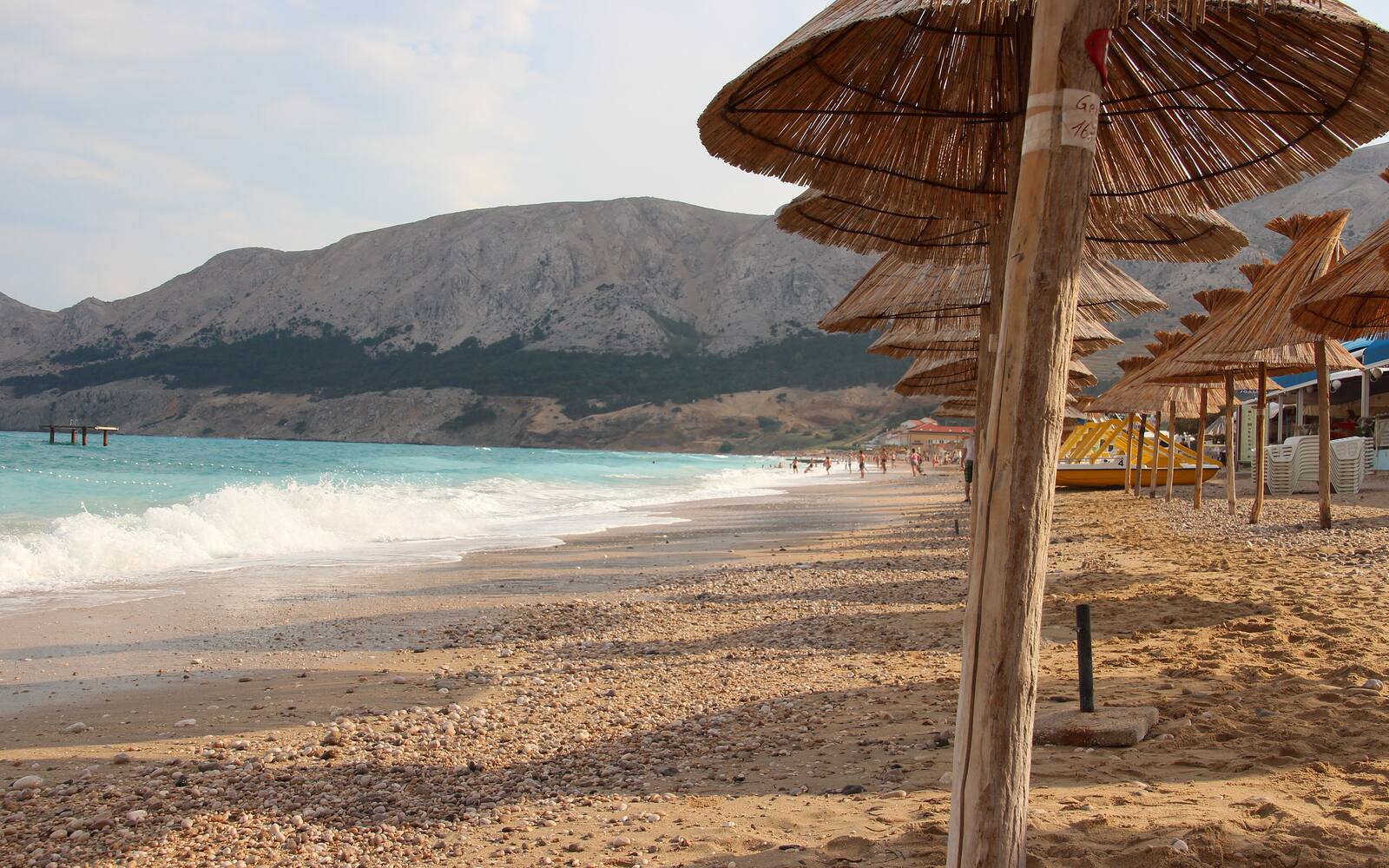 Kiezelstrand van Baška op het eiland Krk met strandparasols van riet en kale bergen op de achtergrond