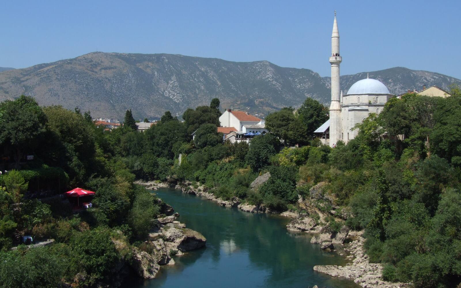 De Neretva stroomt door een groene vallei in Mostar, met een moskee met minaret en kale bergen erachter